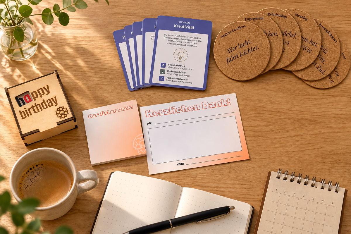 Arbeitstisch von oben mit Stärkenatlas-Karten, PERMA-Korkuntersetzern, Dankzetteln, einer Happy-Birthday-Holzbox, Notizbuch, Kalender und Kaffeetasse. Werkzeuge für Positive Leadership im Alltag. Bild vollständig mit KI generiert.